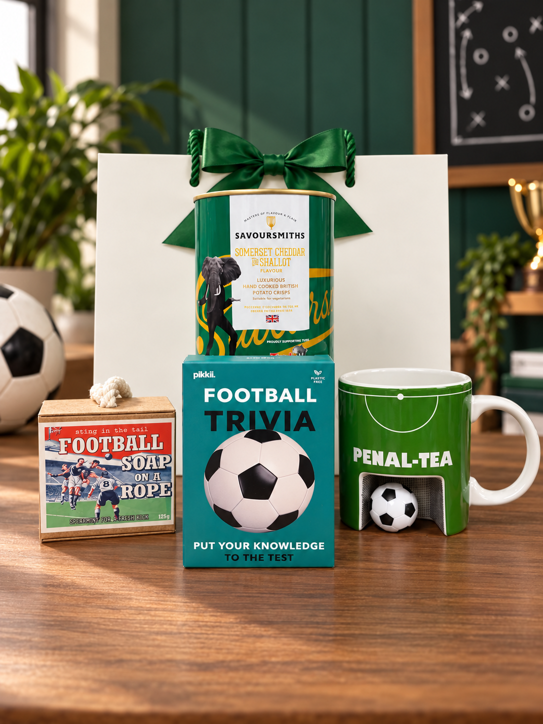 The Football Fan’s Gift Bag on a wooden table, with a Pikkii Football Trivia game, a green Penal-Tea mug with a goal and mini football, a Sting in the Tail football soap on a rope and a tube of Savoursmiths Somerset Cheddar and Shallot crisps, in front of a white gift bag tied with a green satin ribbon.