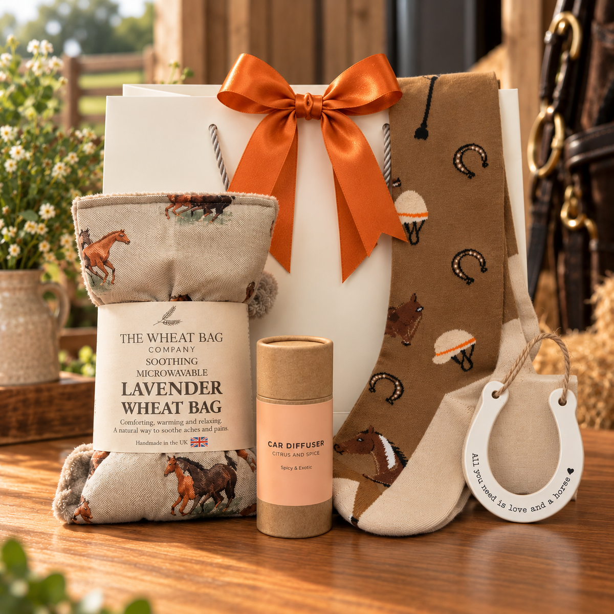 The Horse Lover’s Gift Bag on a wooden table, with a The Wheat Bag Company horse print lavender wheat bag, a pair of horse themed socks, a citrus and spice car diffuser and a ceramic horseshoe reading “All you need is love and a horse”, in front of a white gift bag tied with an orange satin ribbon.