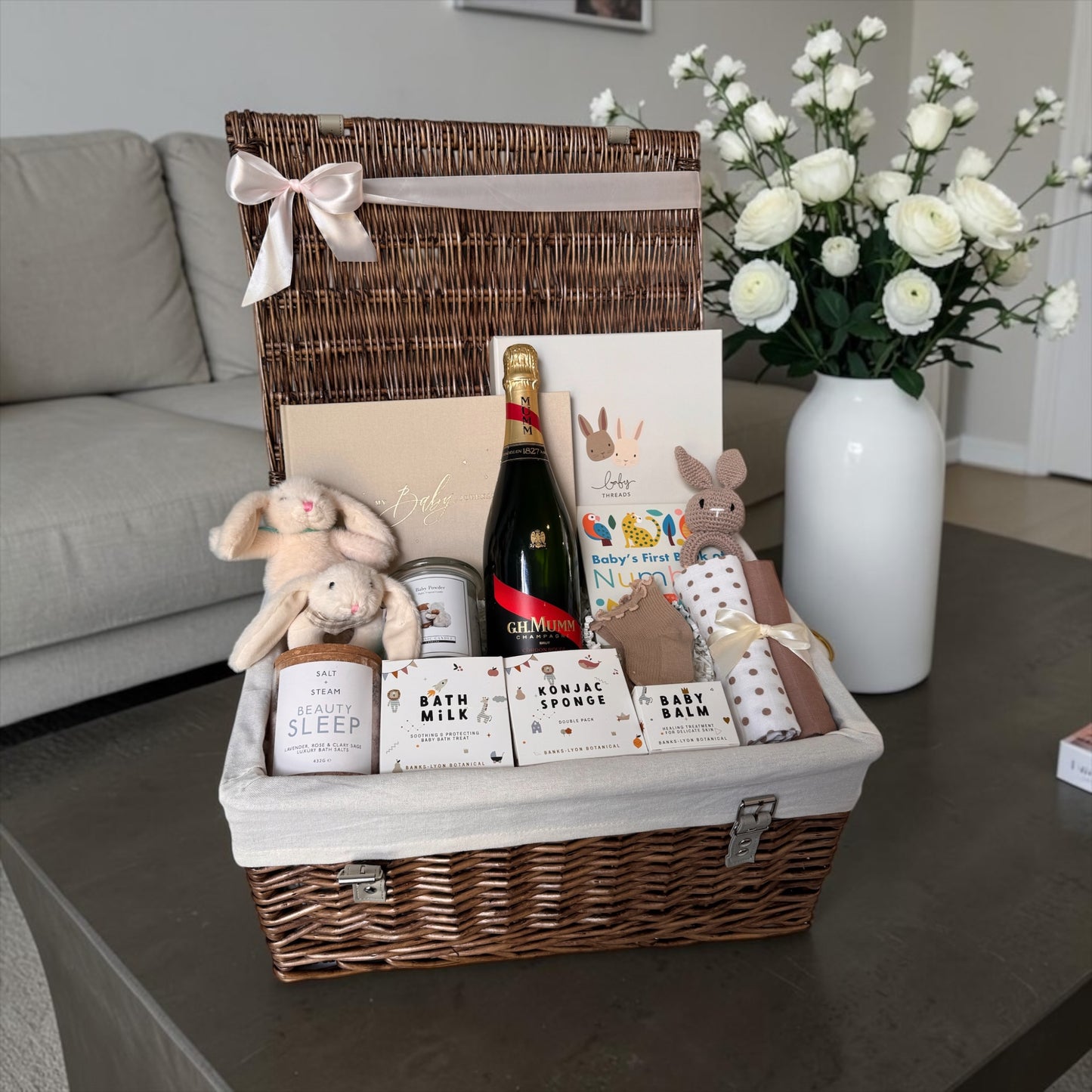 New baby gift hamper with champagne for mum, blush and gold baby journal, teddy in gift box, bunny toys, baby essentials, bath products, and soft accessories in neutral, pink, and baby blue styles.