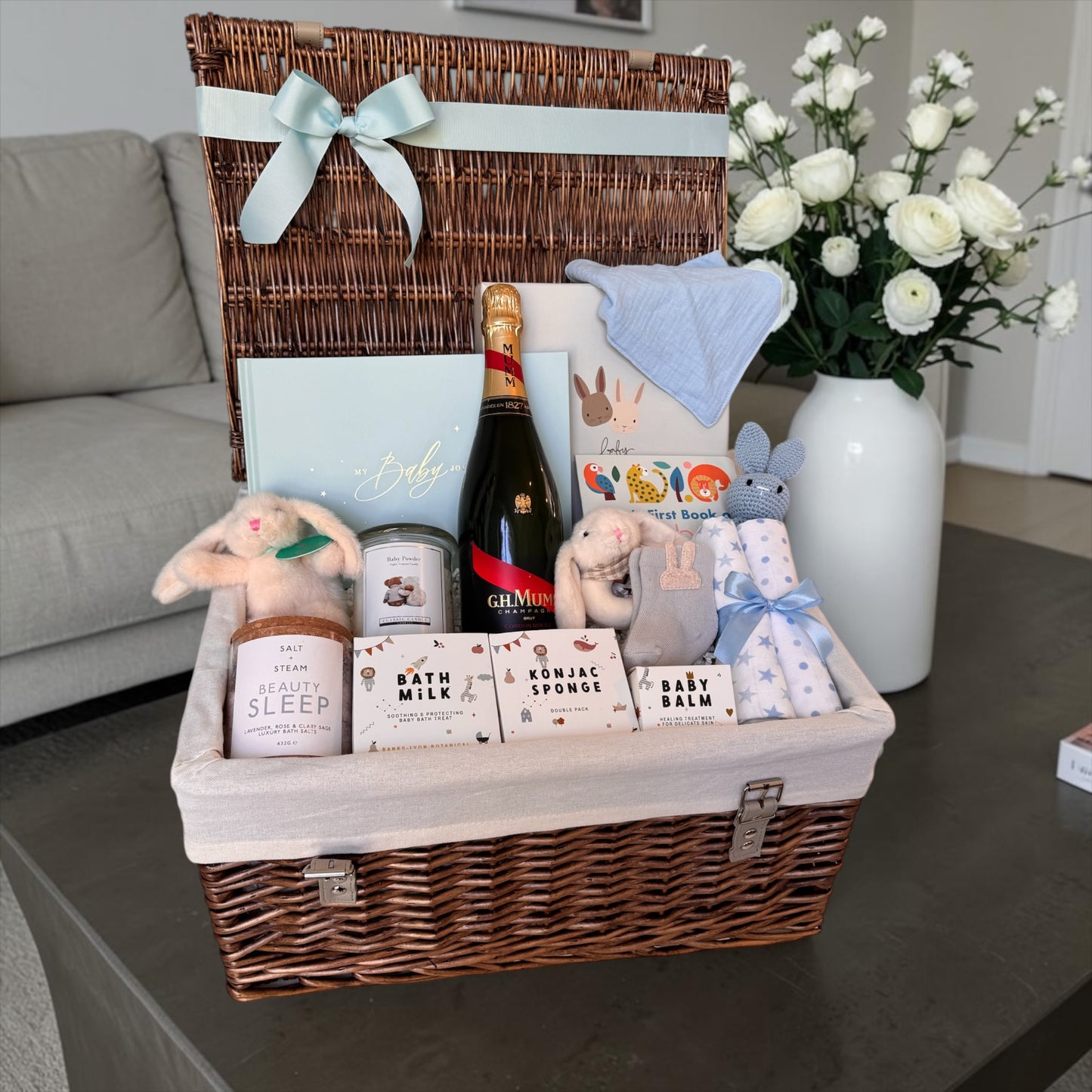 New baby gift hamper with champagne for mum, blush and gold baby journal, teddy in gift box, bunny toys, baby essentials, bath products, and soft accessories in neutral, pink, and baby blue styles.