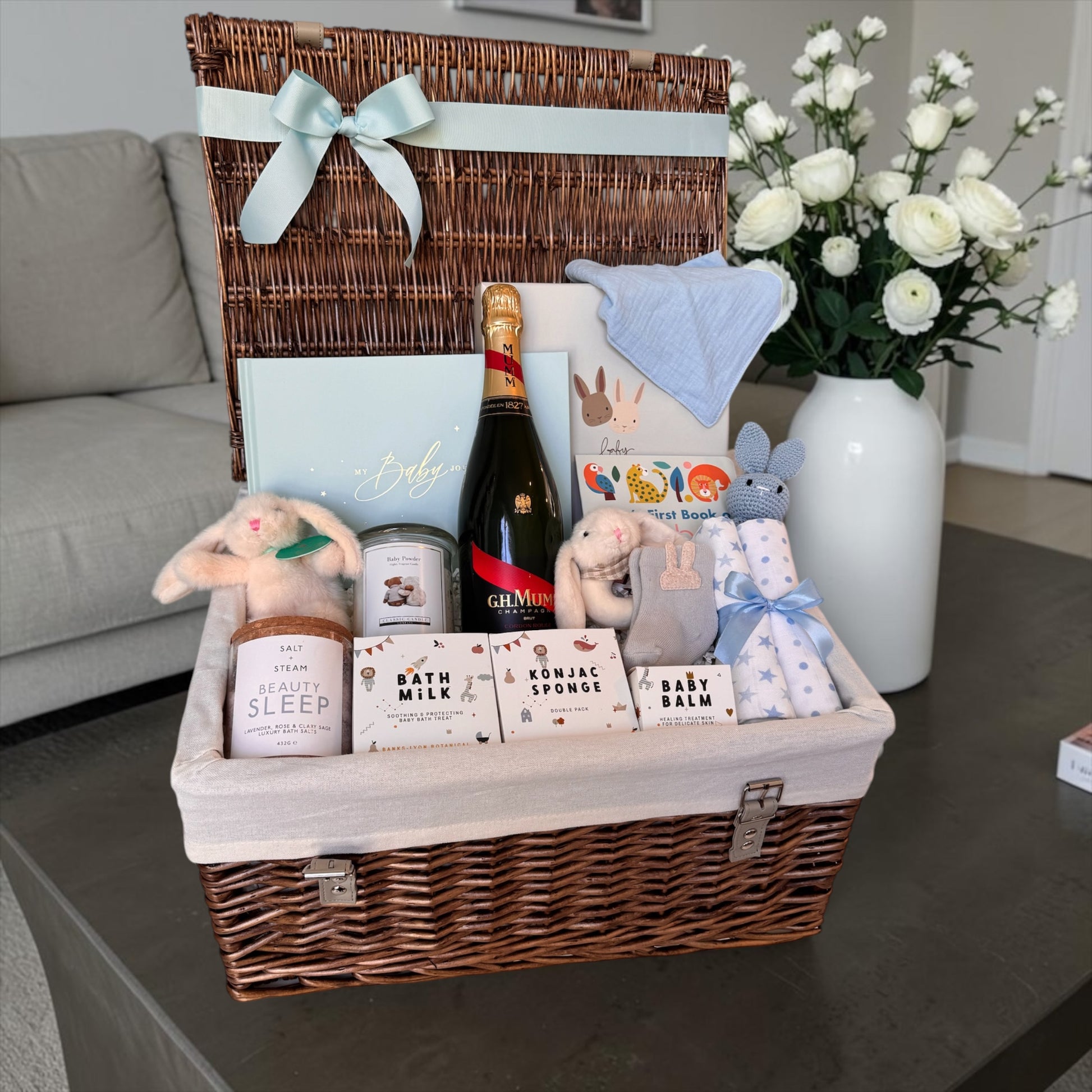 New baby gift hamper with champagne for mum, blush and gold baby journal, teddy in gift box, bunny toys, baby essentials, bath products, and soft accessories in neutral, pink, and baby blue styles.