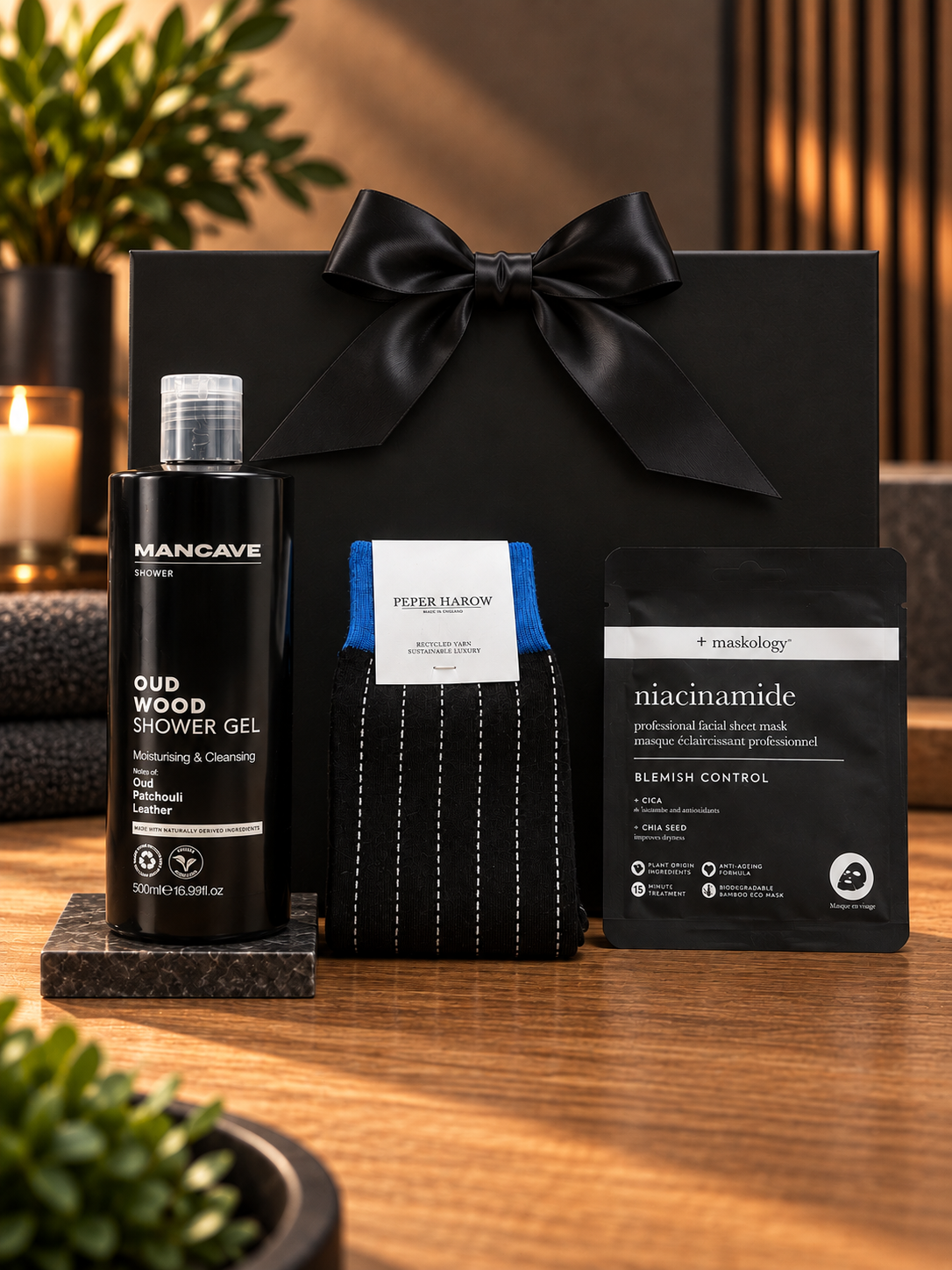 The Oud Wood Pamper Gift Bag on a wooden table, with a 500ml bottle of Mancave Oud Wood shower gel, a pair of black pinstripe Peper Harow socks and a Maskology niacinamide facial sheet mask, in front of a black gift bag tied with a black satin ribbon.