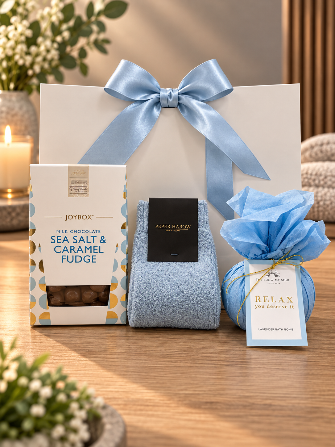 The Relax Gift Bag on a wooden table, with a box of Joybox milk chocolate sea salt and caramel fudge, a pair of soft blue Peper Harow socks and a Relax lavender bath bomb wrapped in blue tissue, in front of a white gift bag tied with a blue satin ribbon.