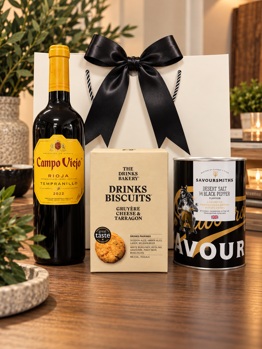 The Rioja & Nibbles Gift Bag with a bottle of Campo Viejo Rioja Tempranillo, a box of The Drinks Bakery Gruyère cheese and tarragon drinks biscuits and a tube of Savoursmiths Desert Salt and Black Pepper crisps, in front of a white gift bag tied with a black satin ribbon.