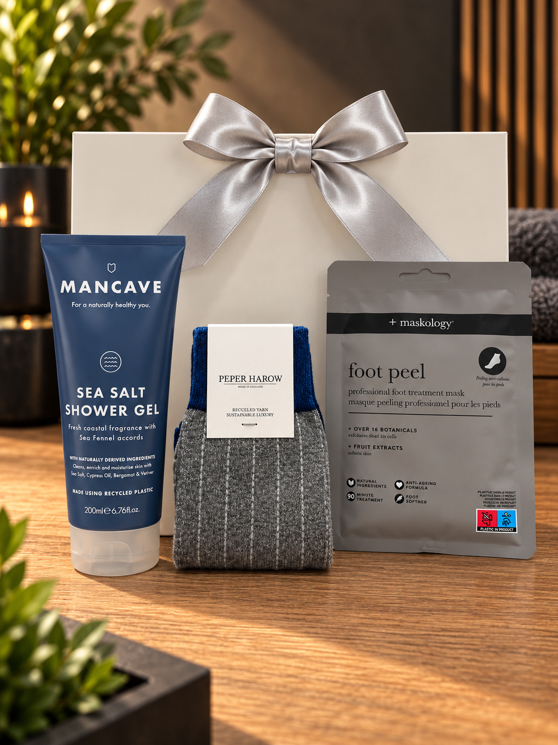 The Sea Salt Pamper Gift Bag on a wooden table, with a tube of Mancave Sea Salt shower gel, a pair of grey pinstripe Peper Harow socks and a Maskology foot peel mask, in front of a white gift bag tied with a silver satin ribbon.