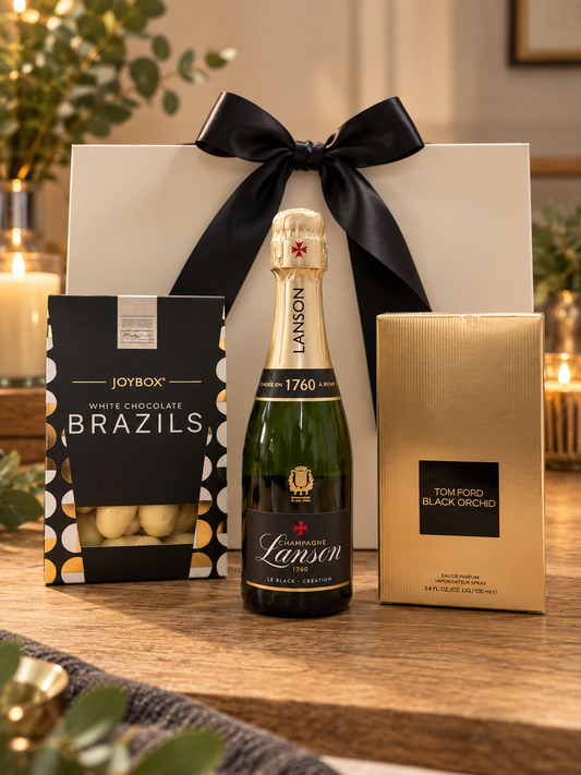 The Tom Ford Black Orchid Gift Bag on a wooden table, with a box of Joybox white chocolate Brazils, a mini bottle of Lanson Champagne and a boxed 100ml bottle of Tom Ford Black Orchid eau de parfum, in front of a white gift bag tied with a black satin ribbon.