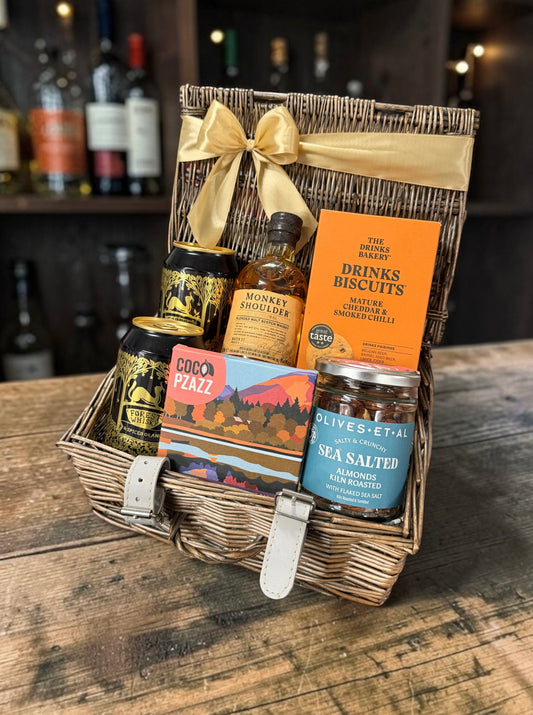 A wicker gift basket with a gold ribbon and bow, filled with assorted gourmet items including a bottle of Monkey Shoulder Scotch, a box of The Drinking Biscuits, Cocoa Pazzz chocolate, and Olives & Etal sea salted olives, set on a rustic wooden table with a blurred bar background.