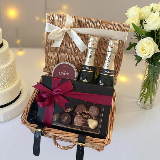 Gift basket with bottles, chocolates, and a card on a table with a cake and flowers in the background.