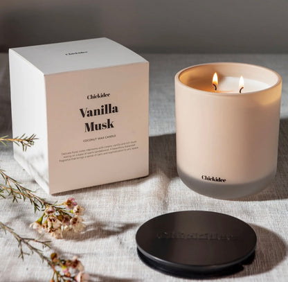 Chicklider Vanilla Musk coconut wax candle shown with its white square box, a lit frosted glass candle jar, and a black lid on a textured linen surface with dried flowers.