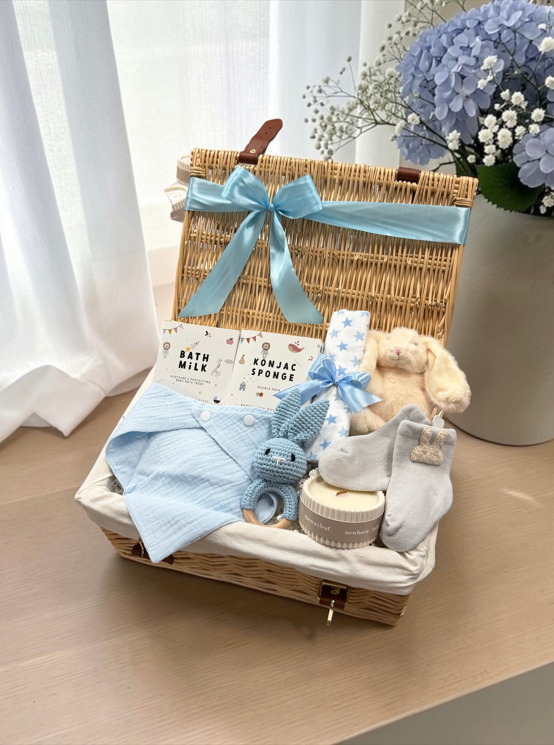 A baby gift basket set in a wicker hamper with a light blue ribbon bow, filled with baby items including a blue striped cloth, a plush bunny, a blue-and-white muslin cloth; a sponge, and socks, displayed on a wooden table with a vase of blue flowers in the background.