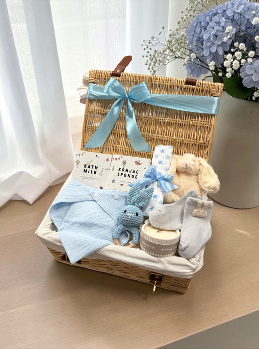 A baby gift basket set in a wicker hamper with a light blue ribbon bow, filled with baby items including a blue striped cloth, a plush bunny, a blue-and-white muslin cloth; a sponge, and socks, displayed on a wooden table with a vase of blue flowers in the background.