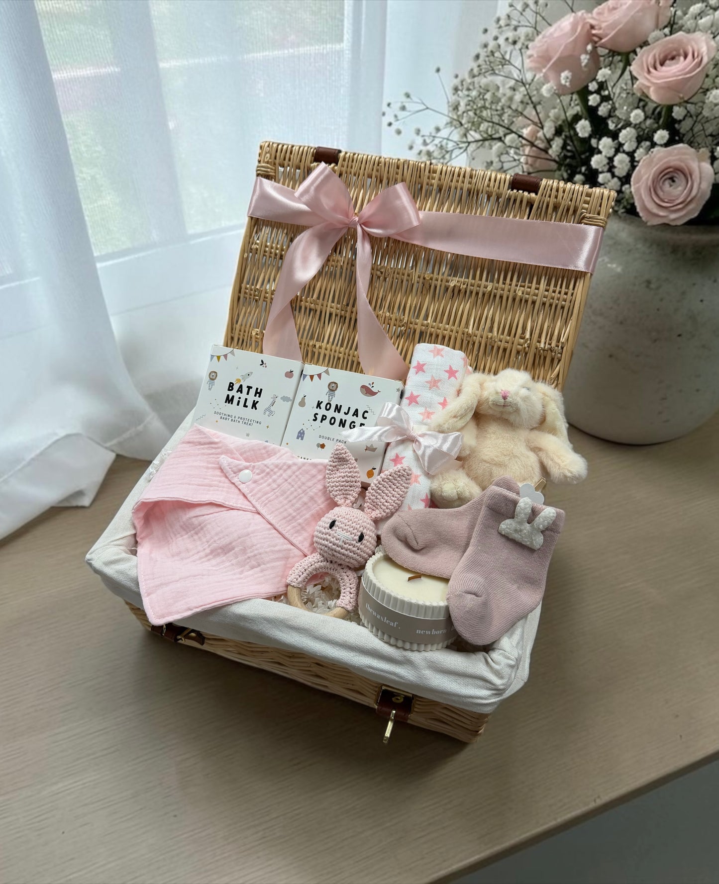 A baby gift basket set on a wooden table, featuring a woven wicker basket with a pink satin bow, containing a pink muslin cloth, bunny-shaped bath and body items, a plush bunny, and pink socks.