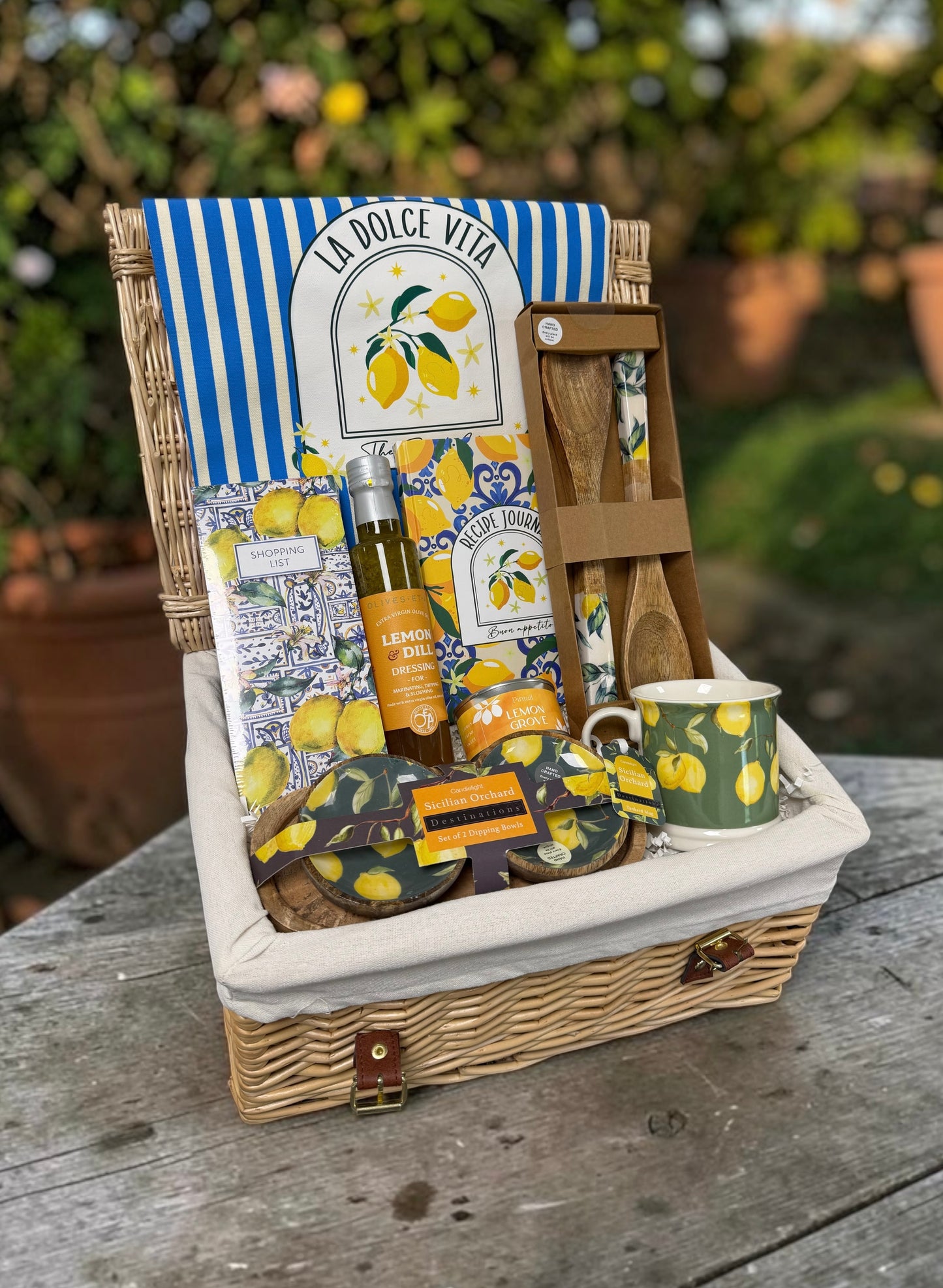 A wicker picnic basket filled with lemon-themed items, including a striped and patterned La Dolce Vita towel, a bottle of olive oil, wooden utensils, a green-and-yellow mug, and small packaged snacks, set on a wooden table outdoors.