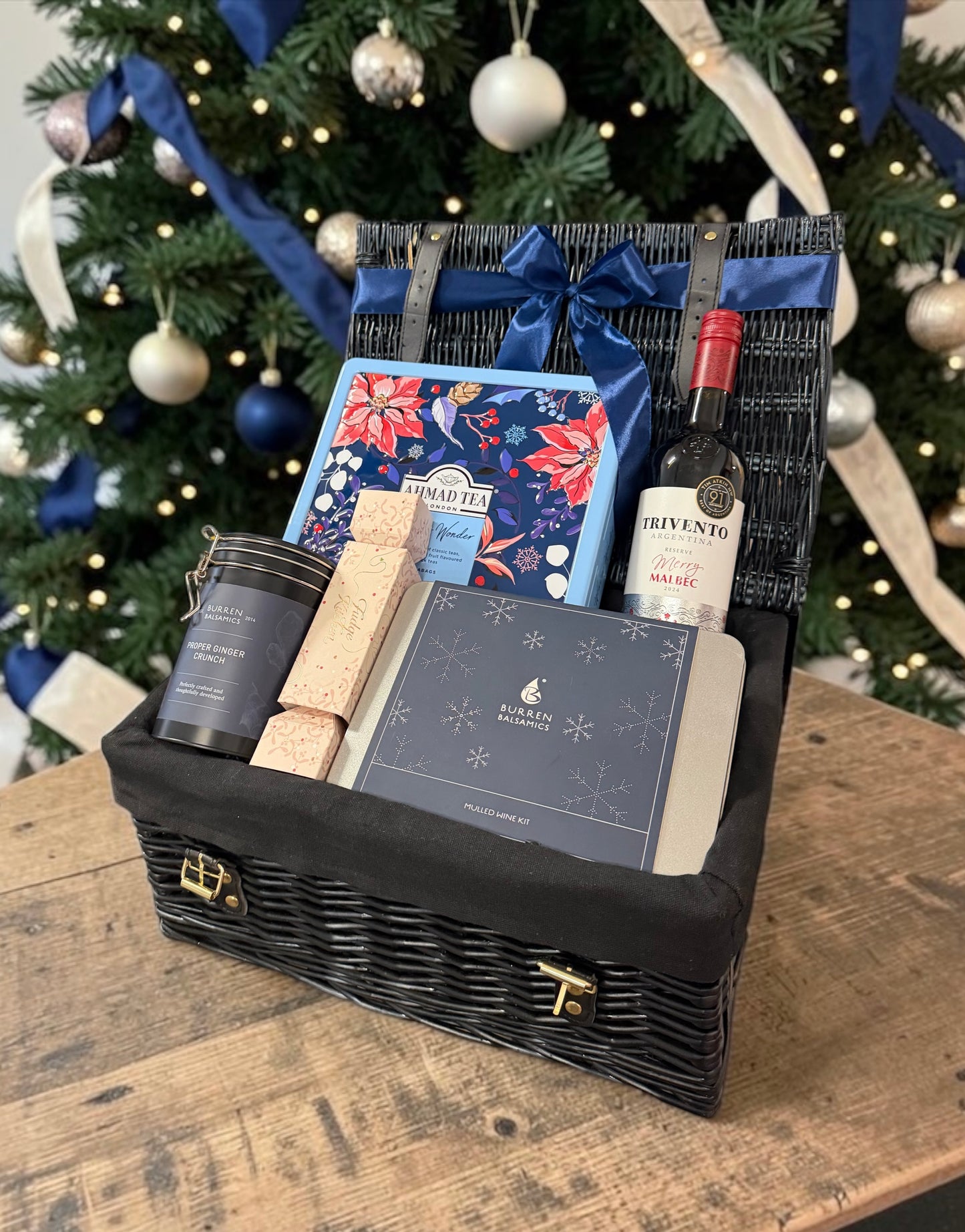 A Christmas-themed gourmet gift hamper in a black wicker basket, featuring a bottle of red wine, a tin of ginger biscuits, a mulled wine kit, and fudge cracker, all arranged with a blue ribbon bow in front of a decorated Christmas tree.