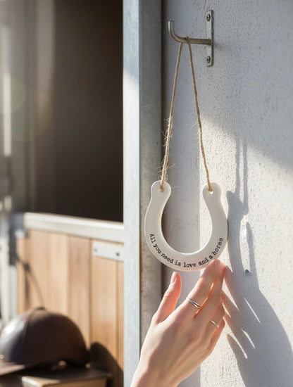 A white horseshoe-shaped wall hanging with a rope loop and printed quote is being held by a person's hand against a sunlit gray wall.