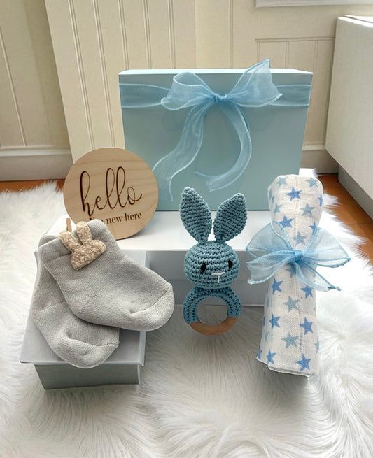 Baby Boy Gift Set featuring soft blue bunny socks, blue bunny rattle, breathable muslin cloth, and “Hello, I’m New Here” plaque for a newborn.