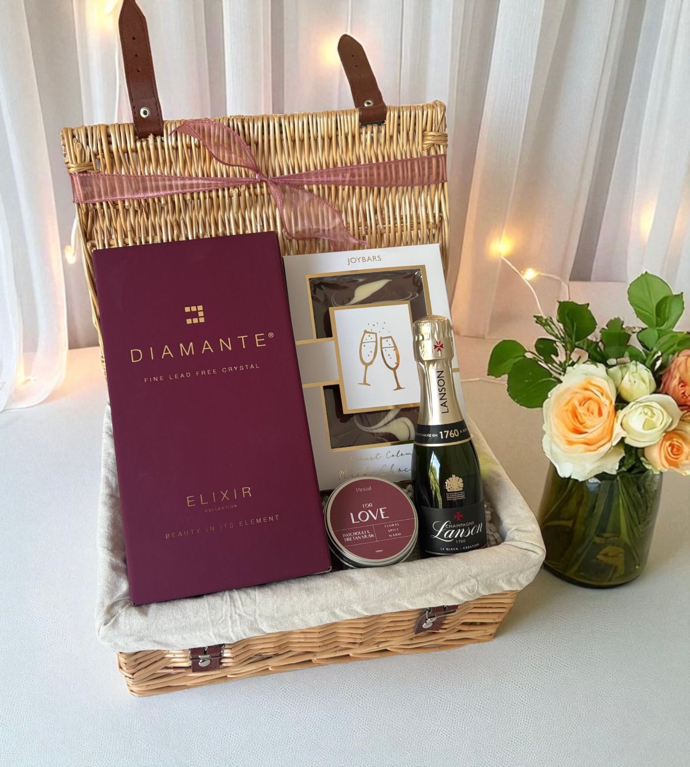 Gift basket with Diamante Elixir, Lanvin chocolate, and flowers on a white surface.