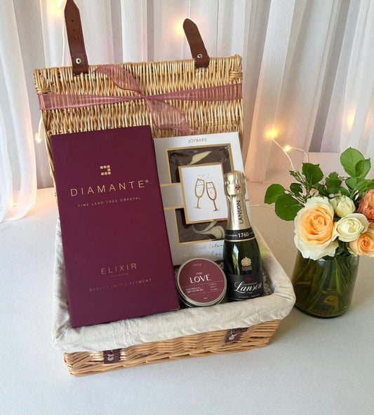 Gift basket with Diamante Elixir, Lanvin chocolate, and flowers on a white surface.