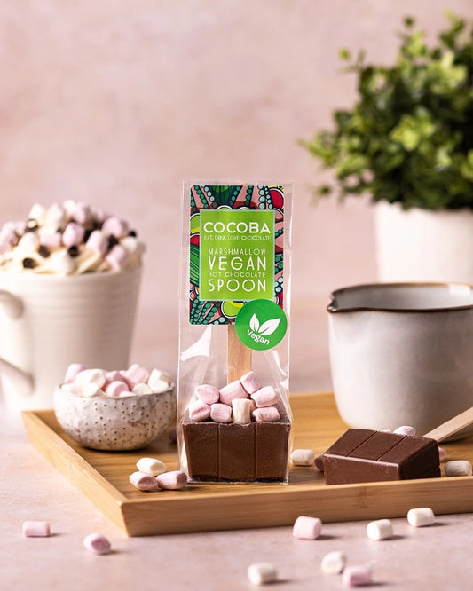 A packaged chocolate marshmallow spoon by Cocoba is displayed on a wooden tray with marshmallows, a mug of whipped cream, and a bowl of marshmallows in a styled setting.