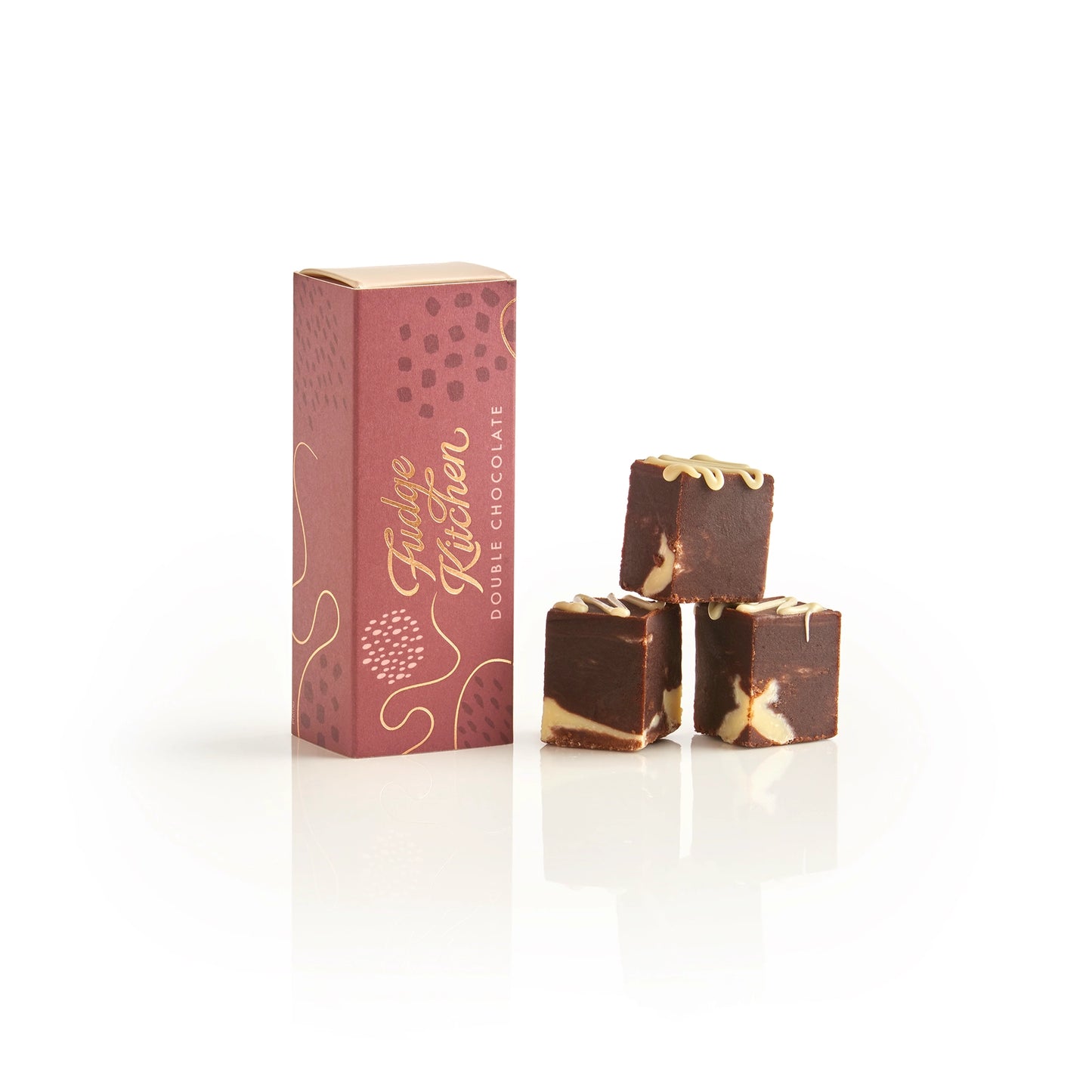 Three pieces of Fudge Kitchen dark chocolate with white drizzle next to its pink packaging box on a white reflective surface.