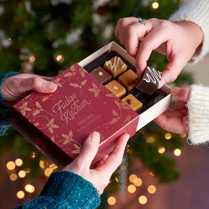 A boxed assortment of chocolates is being opened by multiple hands in front of a blurred Christmas tree with warm lights. The red lid has gold holly and the words 'Fudge Kitchen Festive Chocolate Assortment.