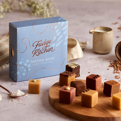 A blue 'Fudge Kitchen' tasting menu box for the Fudge Collection stands on a styled tabletop with assorted chocolates and caramel cubes arranged on a wooden board and beside a small cream pitcher.