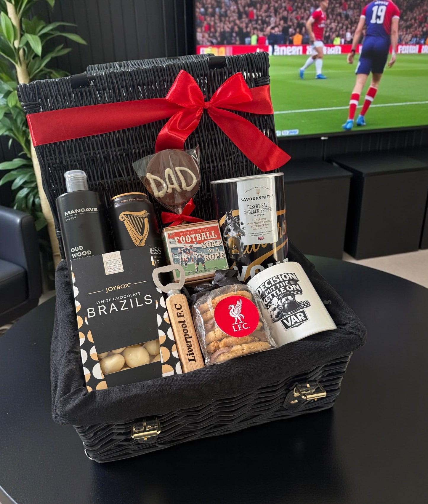 Gift basket with snacks and drinks on a table in front of a television showing a football match.