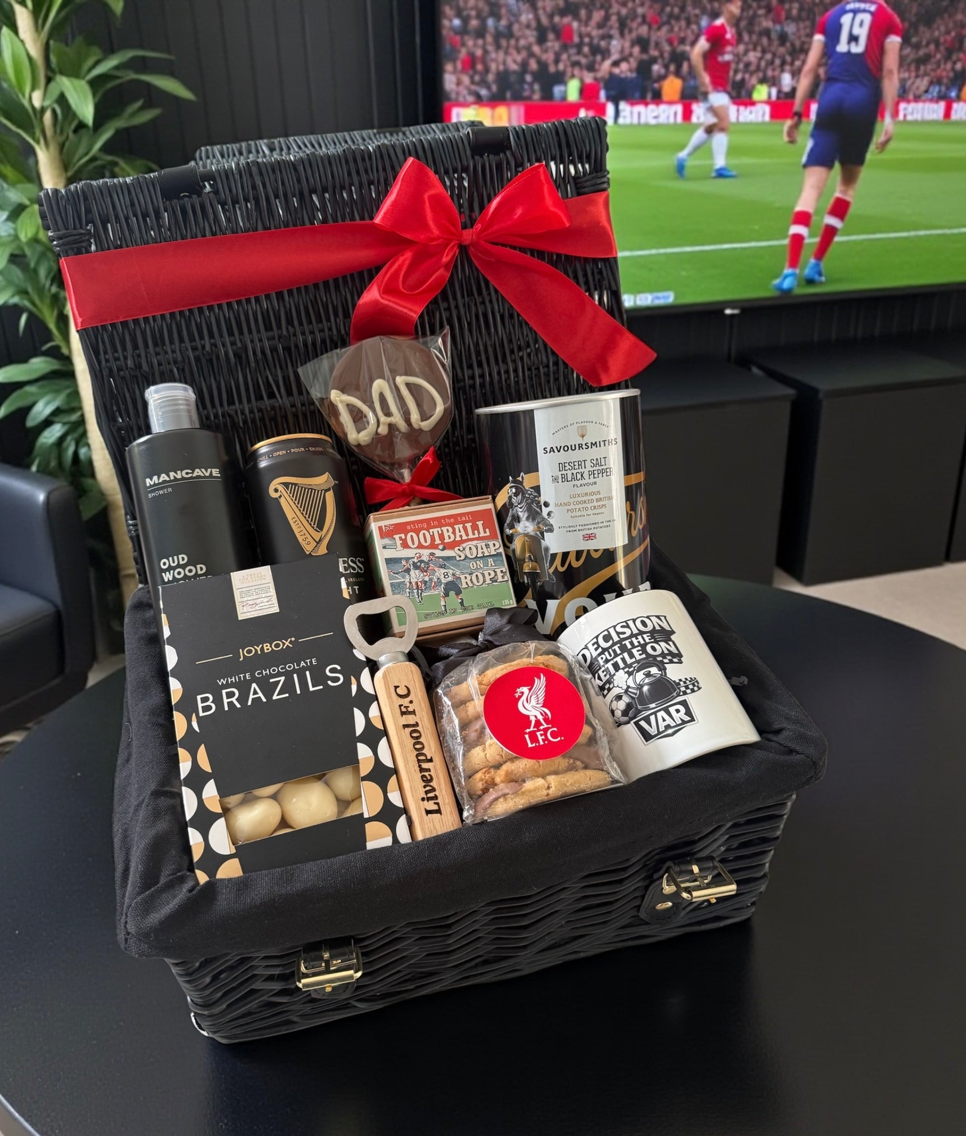 Gift basket with snacks and drinks on a table in front of a television showing a football match.