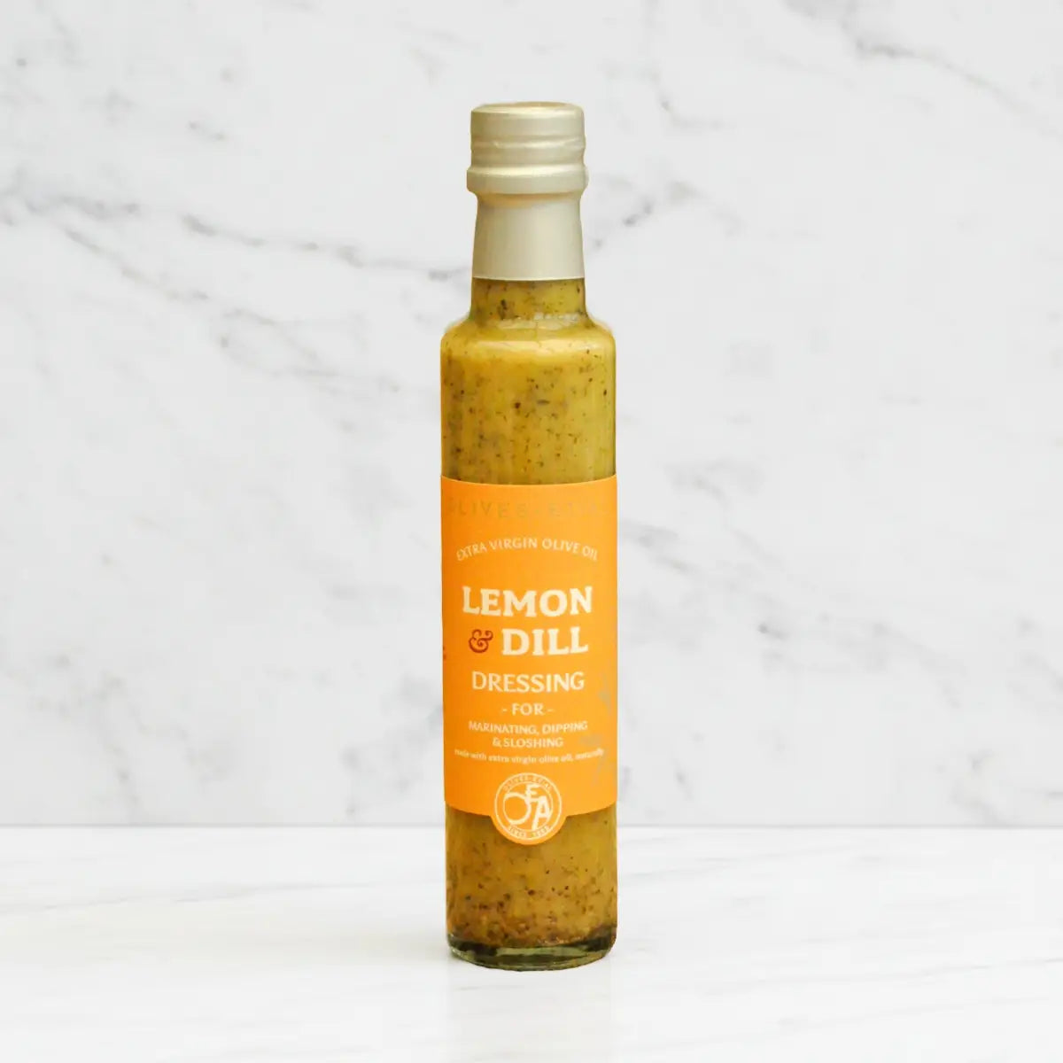 A bottle of FA Lemon & Dill dressing for salad, featuring a bright orange label and a gold cap, set against a white marble background.