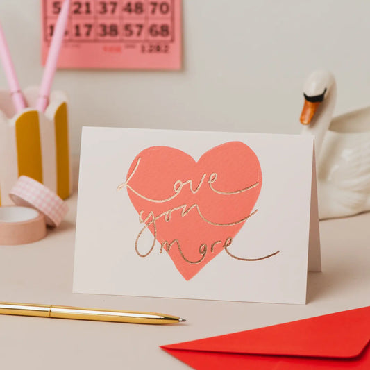 Love You More red heart rose gold foil Valentine's card on a desk.