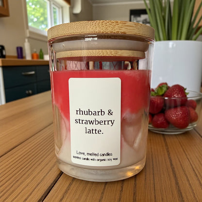 Candle jar labeled 'rhubarb & strawberry latte' on a wooden table with strawberries in the background.