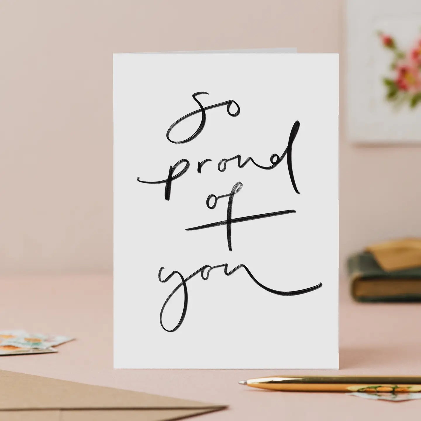 Greeting card with 'So proud of you' text on a desk with stationery items.