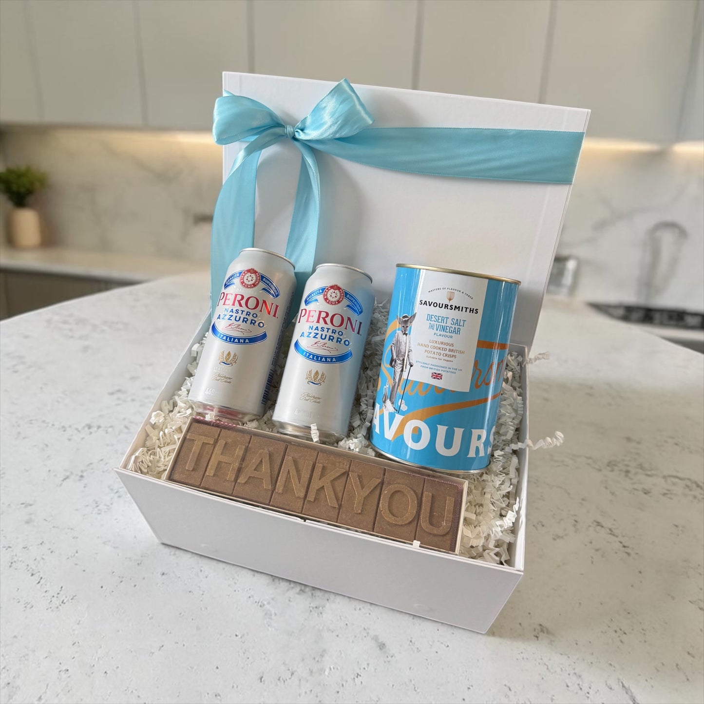 Gift box with Peroni beer cans, a cookie, and a blue ribbon on a kitchen counter.