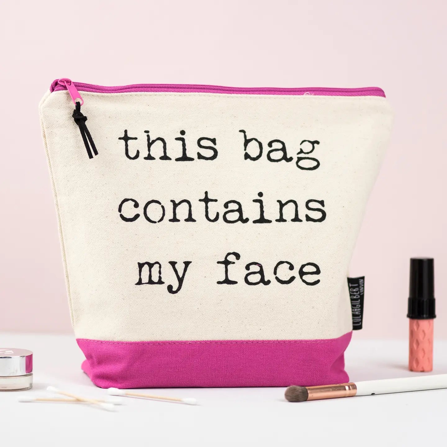 Pink washbag with text "This Bag Contains My Face," crafted from cream canvas with black nylon lining, designed for makeup and toiletries.